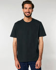 A man with short dark hair and a beard is smiling, wearing a plain black MyNeedsAreSimple BUNDLE X 50 Stanley/Stella Creator 2.0 T-Shirt with free DTG print and dark pants, posed against a neutral background.