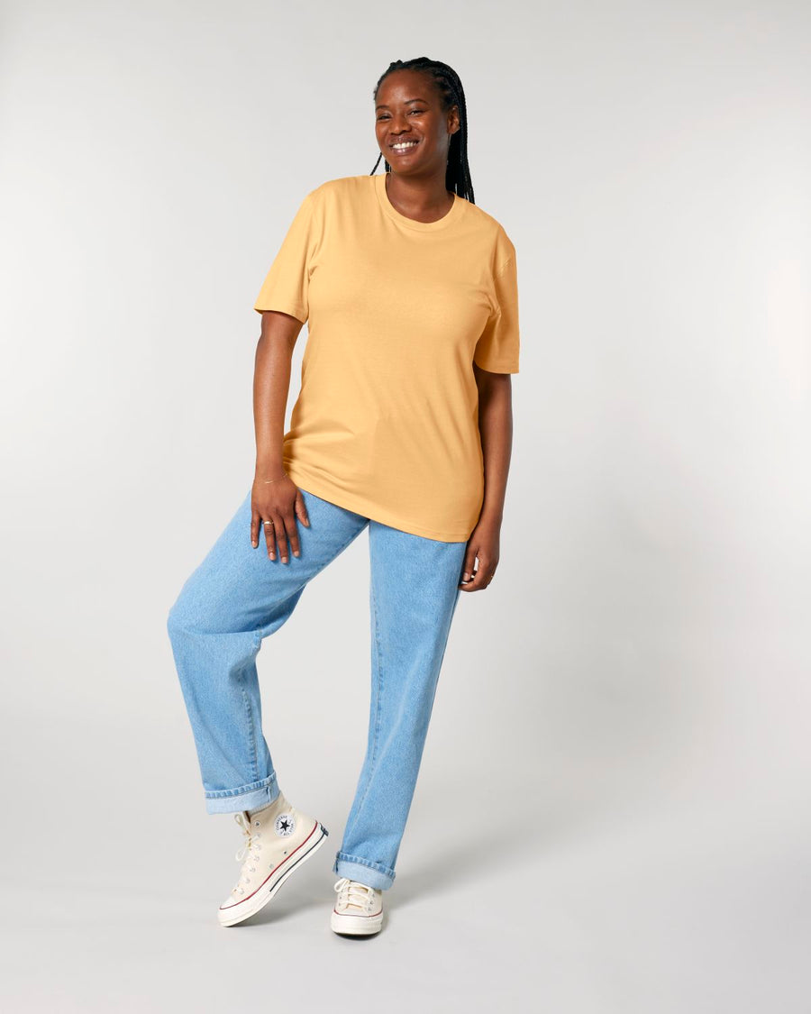 A person standing confidently in a studio, wearing a Stanley/Stella organic cotton mustard yellow STTU169 Creator 2.0 The Iconic unisex t-shirt, blue jeans, and white sneakers.