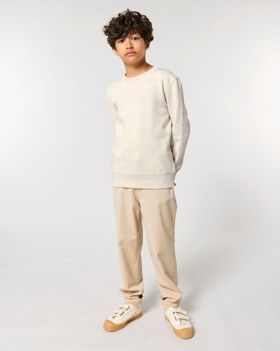A young boy wearing a beige STSK181 Stella/Stella Mini Changer 2.0 The Iconic Kids’ Crew Neck Sweatshirt and tan pants.
