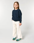 A girl in a blue STSK181 Stella Kids' Crew Neck Sweatshirt and white pants.