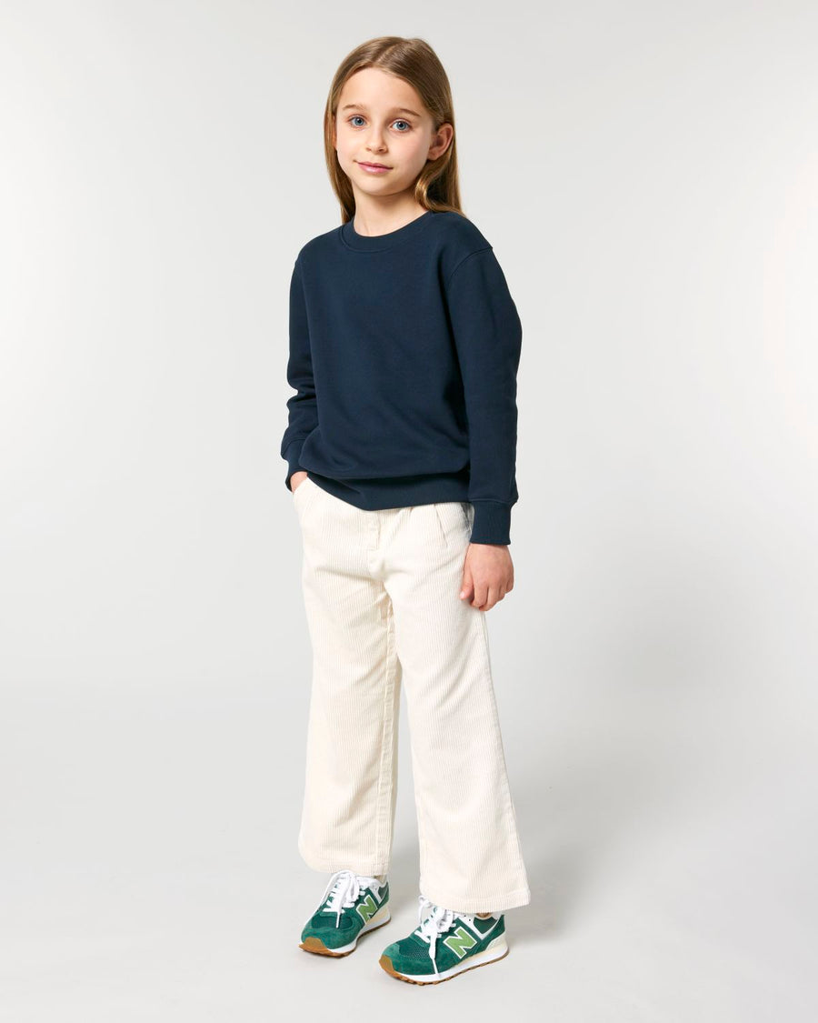 A girl in a blue STSK181 Stella Kids' Crew Neck Sweatshirt and white pants.