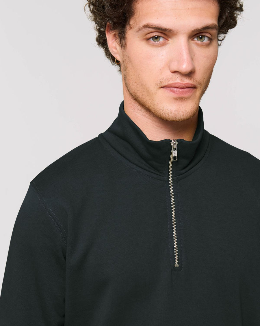 A person with curly hair wears a black Stanley/Stella STSM611 Stanley Trucker Men's Quarter Zip Organic Cotton Sweatshirt, looking at the camera.
