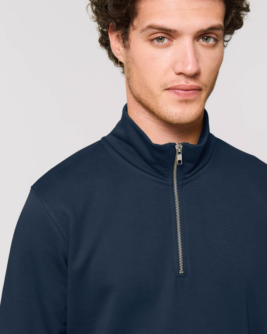 A person with short curly hair is wearing a dark blue Stanley/Stella STSM611 Stanley Trucker Men's Quarter Zip Organic Cotton Sweatshirt and looking at the camera.