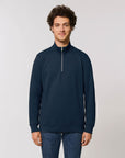 A man with curly hair stands facing the camera, wearing a dark blue Stanley/Stella STSM611 Stanley Trucker Men's Quarter Zip Organic Cotton Sweatshirt, paired with blue jeans against a plain white background.