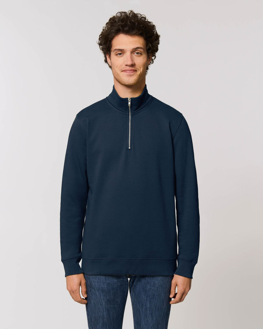 A man with curly hair stands facing the camera, wearing a dark blue Stanley/Stella STSM611 Stanley Trucker Men's Quarter Zip Organic Cotton Sweatshirt, paired with blue jeans against a plain white background.