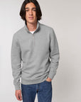 A person with medium-length dark hair wearing a STSM611 Stanley Trucker Men's Quarter Zip Organic Cotton Sweatshirt by Stanley/Stella and blue jeans stands against a plain white background.