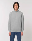 A person with shoulder-length hair is smiling, wearing a Stanley/Stella STSM611 Stanley Trucker Men's Quarter Zip Organic Cotton Sweatshirt, paired with blue jeans. The background is plain and light-colored.