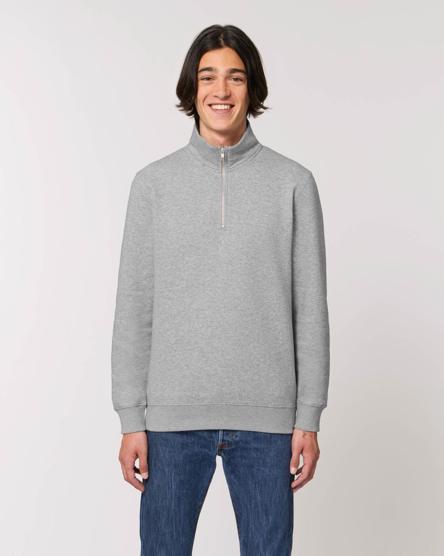 A person with shoulder-length hair is smiling, wearing a Stanley/Stella STSM611 Stanley Trucker Men's Quarter Zip Organic Cotton Sweatshirt, paired with blue jeans. The background is plain and light-colored.