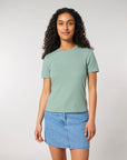 A woman with long curly hair wearing a light green STTW174 Stella Ella Womens Fitted Loose Knit T-Shirt from Stanley/Stella and a denim skirt stands against a plain background.