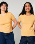 Two women in yellow STTW174 Stella Ella fitted t-shirts and jeans standing side by side, smiling against a gray background.