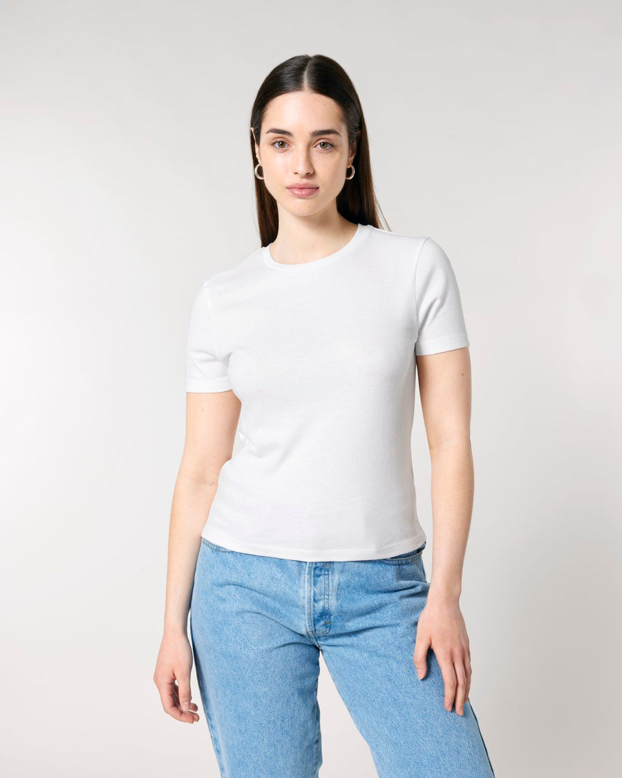 A woman with long, dark hair stands against a neutral background. She wears a plain white **Stanley/Stella STTW174 Stella Ella Womens Fitted Loose Knit T-Shirt** made from organic cotton and blue jeans, with one hand resting casually at her side.