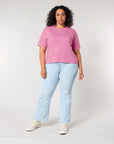Woman standing against a grey background wearing a STTW175 Stella Nova Women's Boxy T-Shirt by Stanley/Stella, blue jeans, and white sneakers.