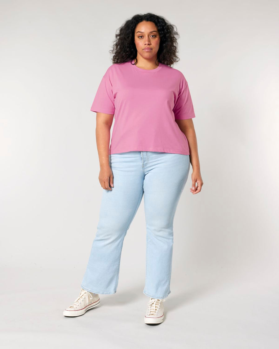 Woman standing against a grey background wearing a STTW175 Stella Nova Women's Boxy T-Shirt by Stanley/Stella, blue jeans, and white sneakers.