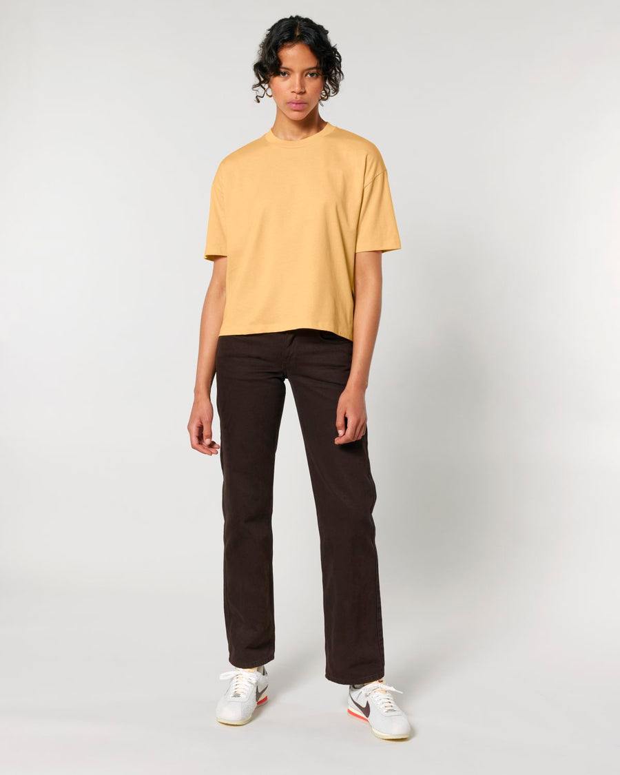 Person standing in a neutral pose wearing a STTW175 Stella Nova Womens Boxy T-Shirt from Stanley/Stella in yellow, dark trousers, and white sneakers.
