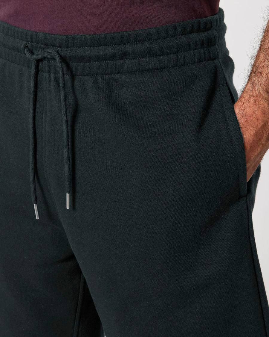 Close-up of a person wearing black STBU186 Stella/Stella Trainer 2.0 The Iconic Mid-light Unisex Jogger Shorts by Stanley/Stella and a dark shirt, with their right hand in their pocket.