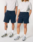 Two people stand side by side wearing matching grey shirts, Stanley/Stella STBU186 Stella/Stella Trainer 2.0 The Iconic Mid-light Unisex Jogger Shorts in navy blue, white socks, and athletic shoes.