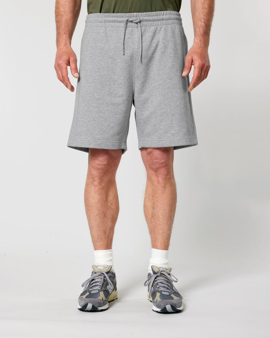 Person wearing a gray T-shirt, Stanley/Stella STBU186 Stella/Stella Trainer 2.0 The Iconic Mid-light Unisex Jogger Shorts made from organic cotton, white socks, and gray sneakers standing against a plain background.