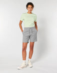 A person stands with hands in pockets, wearing a light green t-shirt, gray unisex jogger shorts made from French terry (STBU186 Stella/Stella Trainer 2.0 The Iconic Mid-light Unisex Jogger Shorts by Stanley/Stella), white socks, and white sneakers. The background is plain and light.