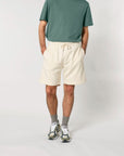 A person stands wearing a green t-shirt, Stanley/Stella STBU186 Stella/Stella Trainer 2.0 The Iconic Mid-light Unisex Jogger Shorts made from organic cotton with hands in pockets, gray socks, and green-and-white sneakers. The background is plain white.
