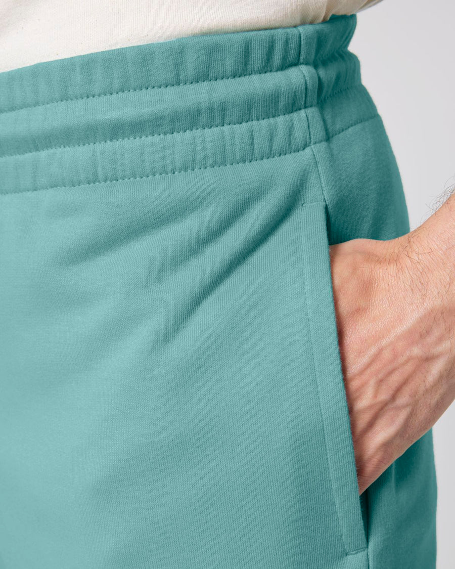 Close-up of a person wearing STBU186 Stella/Stella Trainer 2.0 The Iconic Mid-light Unisex Jogger Shorts by Stanley/Stella with their hand inserted into the pocket. The waistband and pocket seam are clearly visible, showcasing the soft French terry fabric crafted from organic cotton.