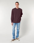 Man standing against a white background wearing a maroon STSU178 Stella crew neck sweatshirt made of organic cotton, blue jeans, and tan sneakers.