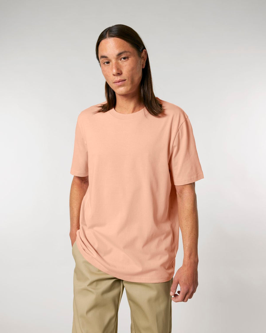 Man standing against a plain background, wearing a Stanley/Stella Creator 2.0 The Iconic Unisex T-Shirt in organic peach color and khaki trousers.
