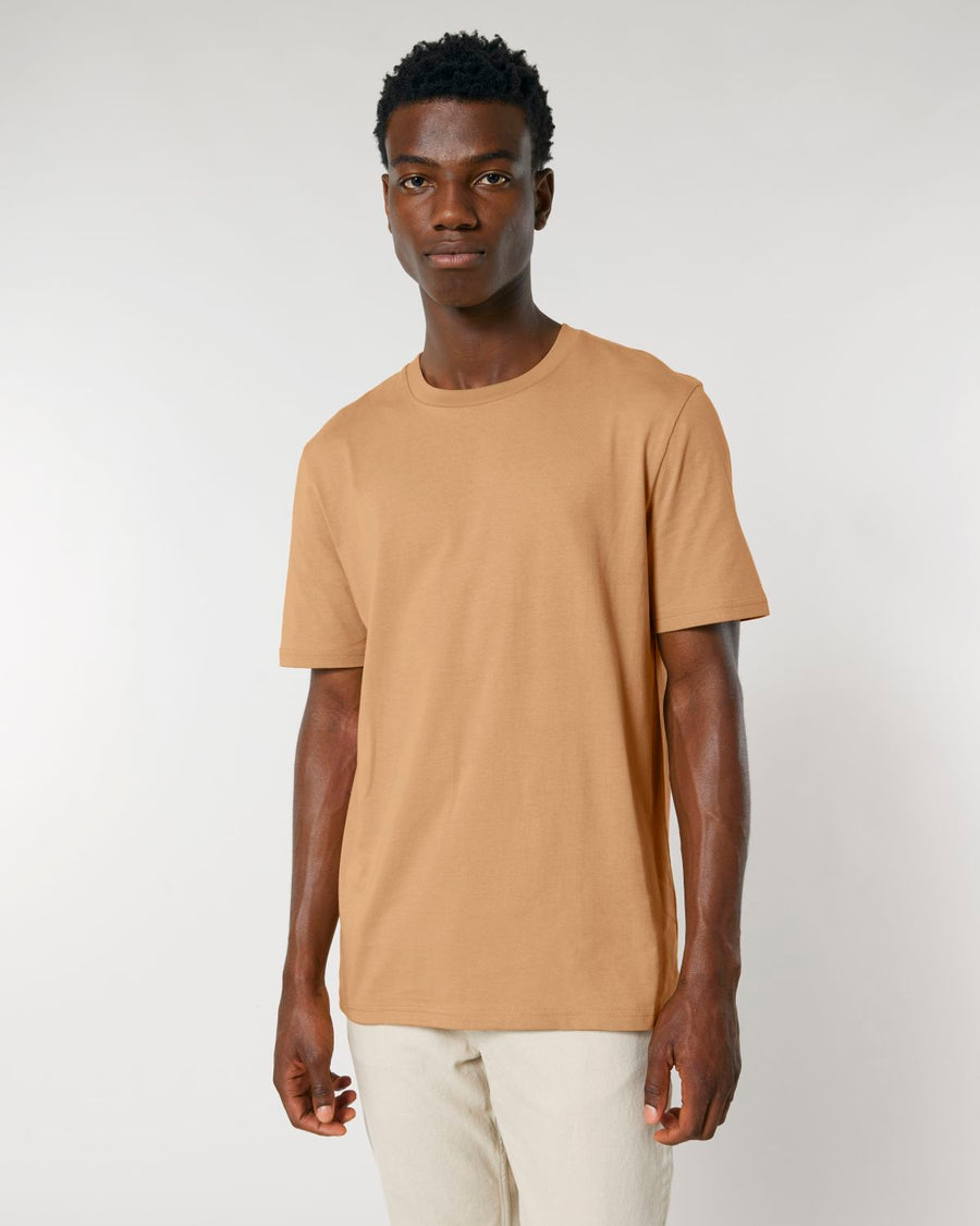 Man in a plain brown Stanley/Stella Creator 2.0 The Iconic Unisex T-Shirt and off-white trousers standing against a neutral background.
