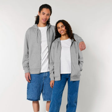 Two individuals stand together, both wearing grey Stanley/Stella STSU179 Stella/Stella Cultivator 2.0 The Iconic Unisex Zip-thru Hoodie Sweatshirts made from brushed fleece and organic cotton, white t-shirts, and blue jeans. One has an arm around the other's shoulder. They are smiling in front of a plain background.