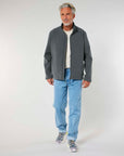 A senior man models casual fashion, standing in a windproof STJM167 Stanley/Stella Navigator Men's Non-Hooded Softshell jacket, blue jeans, and sneakers against a plain background from MyNeedsAreSimple.