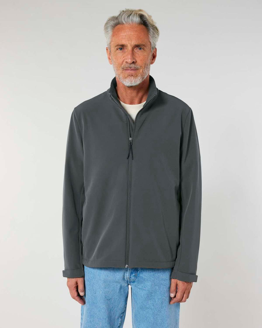 An older man with gray hair, wearing a MyNeedsAreSimple STJM167 Stanley/Stella Navigator Men's Non-Hooded Softshell jacket and light blue jeans, stands facing the camera against a neutral background.