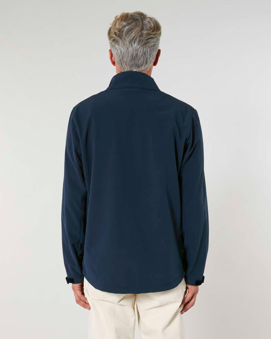 A man standing with his back to the camera, wearing a dark navy MyNeedsAreSimple STJM167 Navigator Men's Non-Hooded Softshell jacket and light beige pants, against a neutral background.
