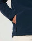 Close-up of a person in a navy, water repellent STJM167 Stanley/Stella Navigator Men's Non-Hooded Softshell hoodie with hand partially inserted into a side zipper pocket, paired with light-colored pants.
