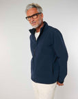 Sentence with brand name and product: A mature man with gray hair, wearing glasses, a blue MyNeedsAreSimple STJM167 Stanley/Stella Navigator Men's Non-Hooded Softshell jacket, and white pants, standing and smiling against a light gray background.
