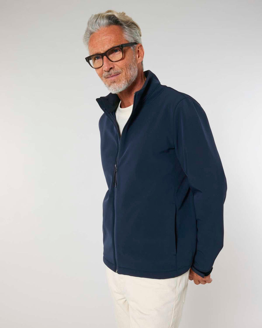 Sentence with brand name and product: A mature man with gray hair, wearing glasses, a blue MyNeedsAreSimple STJM167 Stanley/Stella Navigator Men's Non-Hooded Softshell jacket, and white pants, standing and smiling against a light gray background.