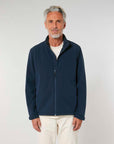 A mature man with gray hair wearing a navy blue MyNeedsAreSimple STJM167 Stanley/Stella Navigator Men's Non-Hooded Softshell zip-up jacket and beige pants stands against a plain light background.