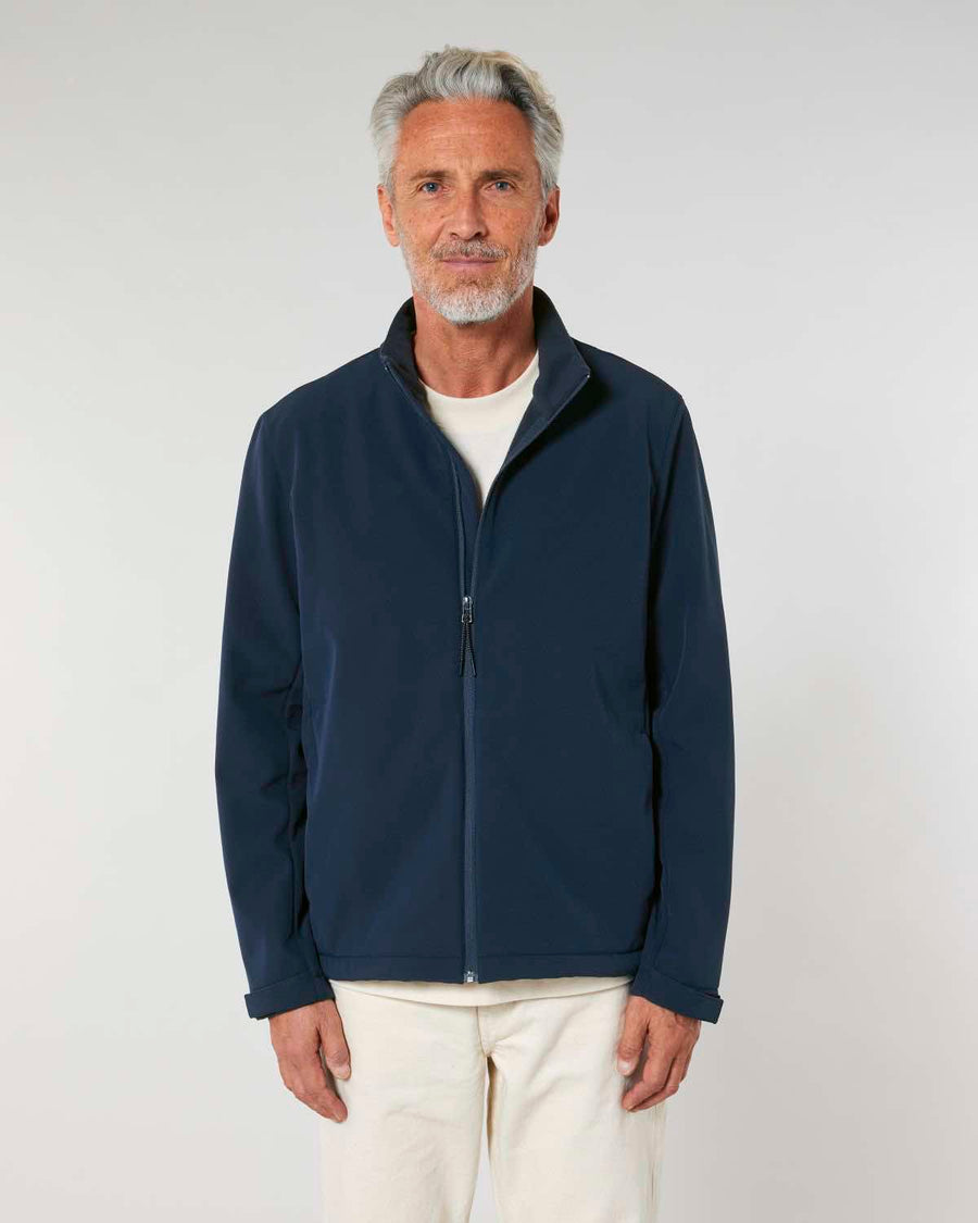 A mature man with gray hair wearing a navy blue MyNeedsAreSimple STJM167 Stanley/Stella Navigator Men's Non-Hooded Softshell zip-up jacket and beige pants stands against a plain light background.