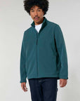 A man with curly hair wearing a STJM167 Stanley/Stella Navigator Men's Non-Hooded Softshell jacket in teal, white t-shirt, and navy trousers stands against a light background.