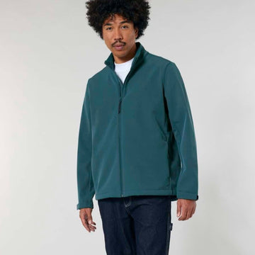A man with curly hair wearing a STJM167 Stanley/Stella Navigator Men's Non-Hooded Softshell jacket in teal, white t-shirt, and navy trousers stands against a light background.