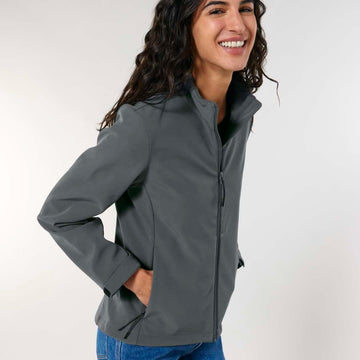 Woman smiling, wearing a MyNeedsAreSimple STJW166 Stanley/Stella Navigator Women’s Non-Hooded Softshell zip-up jacket and blue jeans, standing with one hand on her hip against a light gray background.