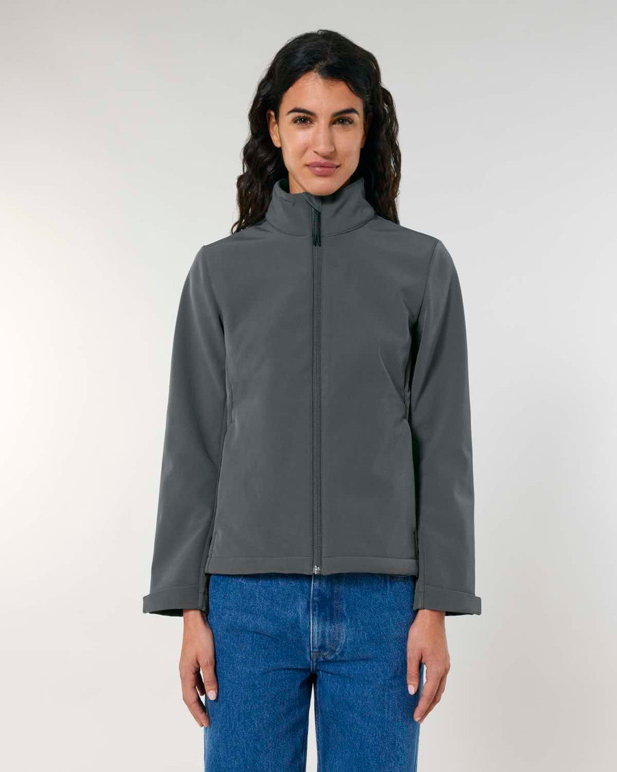 Woman in a grey zip-up STJW166 Stanley/Stella Navigator Women’s Non-Hooded Softshell jacket and blue jeans standing against a light background, looking directly at the camera.
