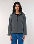 A woman in a MyNeedsAreSimple STJW166 Stanley/Stella Navigator Women’s Non-Hooded Softshell jacket and blue jeans stands facing the camera with a neutral expression.