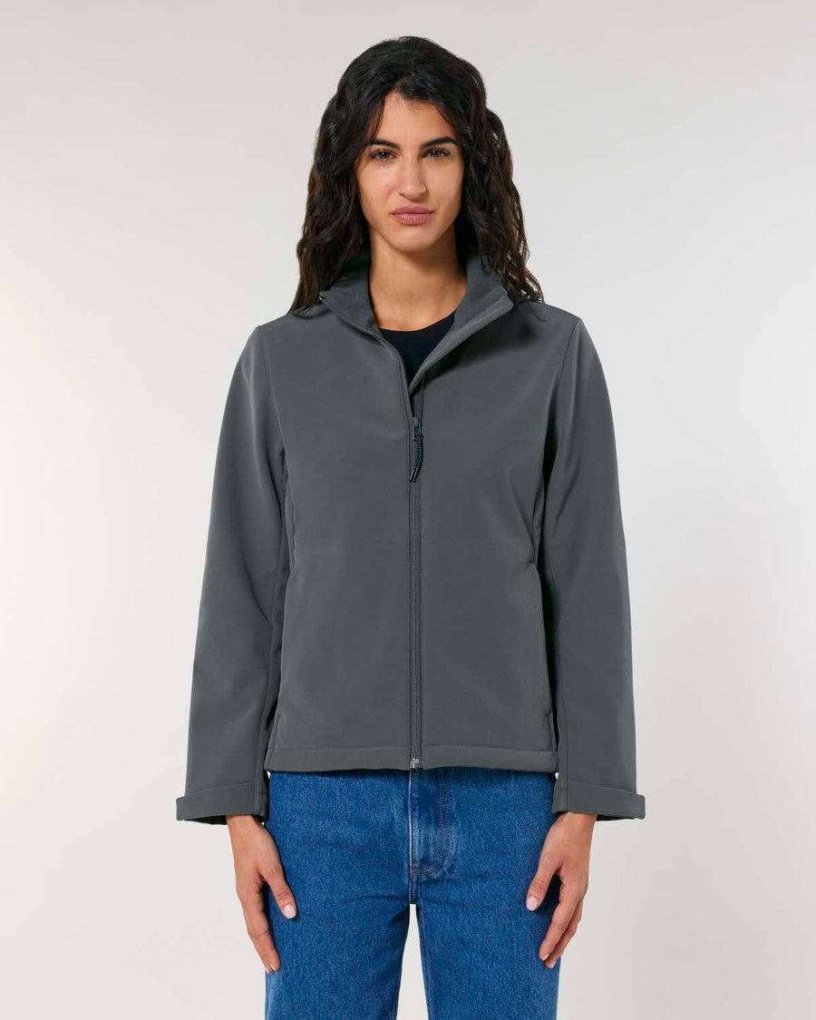 A woman in a MyNeedsAreSimple STJW166 Stanley/Stella Navigator Women’s Non-Hooded Softshell jacket and blue jeans stands facing the camera with a neutral expression.