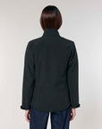 Rear view of a woman wearing a black MyNeedsAreSimple STJW166 Stanley/Stella Navigator Women’s Non-Hooded Softshell and blue jeans against a neutral background.