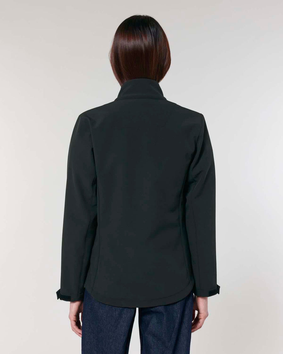 Rear view of a woman wearing a black MyNeedsAreSimple STJW166 Stanley/Stella Navigator Women’s Non-Hooded Softshell and blue jeans against a neutral background.