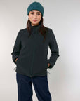Woman wearing a MyNeedsAreSimple STJW166 Stanley/Stella Navigator Women’s Non-Hooded Softshell jacket and blue beanie standing against a light background, hands in pockets, looking at the camera.