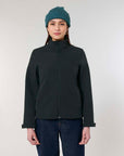 Woman wearing a dark, water-repellent STJW166 Stanley/Stella Navigator Women’s Non-Hooded Softshell jacket and blue beanie, standing against a light gray background.