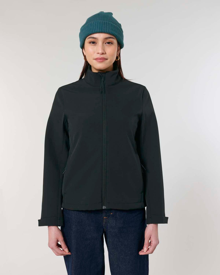 Woman wearing a dark, water-repellent STJW166 Stanley/Stella Navigator Women’s Non-Hooded Softshell jacket and blue beanie, standing against a light gray background.