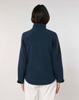 A woman facing away from the camera, wearing a dark blue MyNeedsAreSimple STJW166 Stanley/Stella Navigator Women’s Non-Hooded Softshell blazer and white pants, against a light gray background.