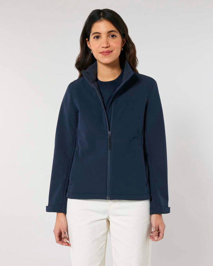 A woman wearing a MyNeedsAreSimple STJW166 Stanley/Stella Navigator Women’s Non-Hooded Softshell jacket and white pants, standing against a light gray background, smiling gently at the camera.