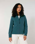 A woman in a breathable, teal STJW166 Stanley/Stella Navigator Women's Non-Hooded Softshell jacket by MyNeedsAreSimple and white pants standing against a plain background.